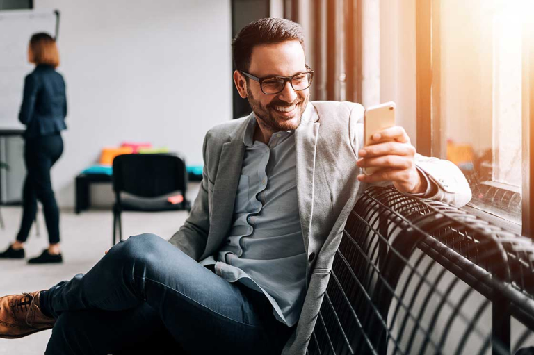 man with glasses wearing sport coat sitting on bench smiling and looking at mobile phone