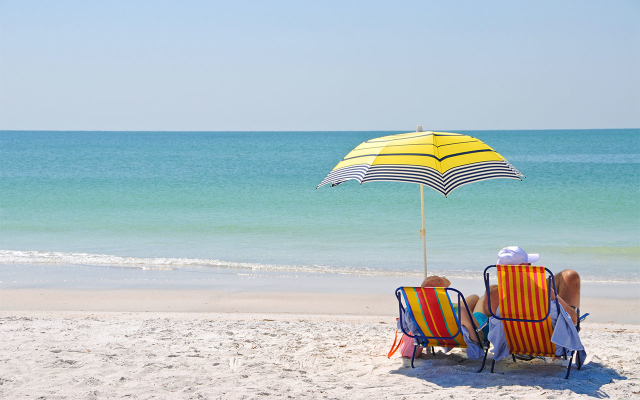 Two people in beach chairs with an umbrella sitting on the beach, looking at the ocean.