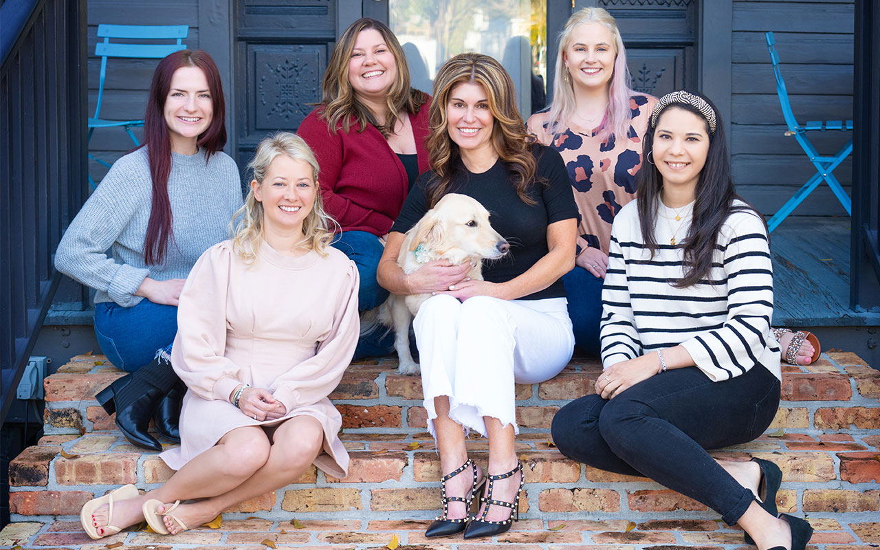 Six women and a dog sitting on stairs in front of a door.