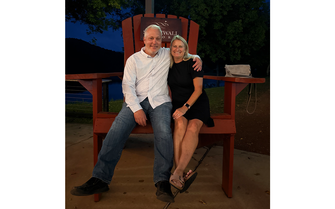 A man and woman sitting next to each other in an oversized wooden chair.
