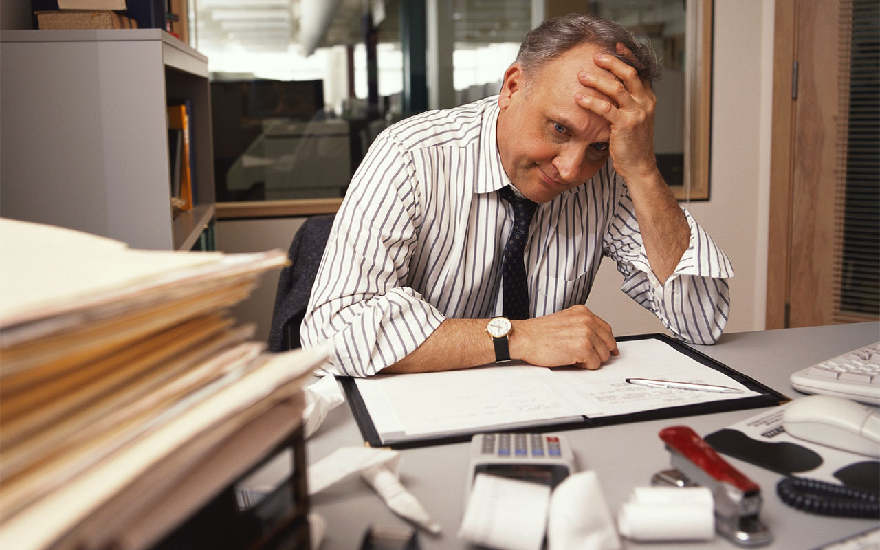 A businessman sitting at a desk with papers and other items all over with his hand on his head.