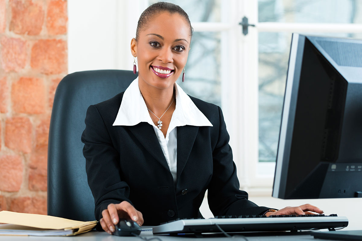 Woman in a black suit sitting at a computer smiling.