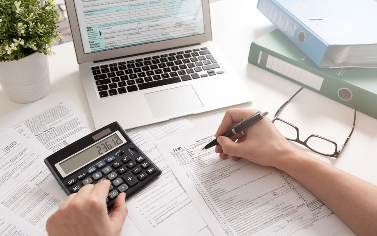 Calculator, laptop, papers, glasses and binders on a desk.
