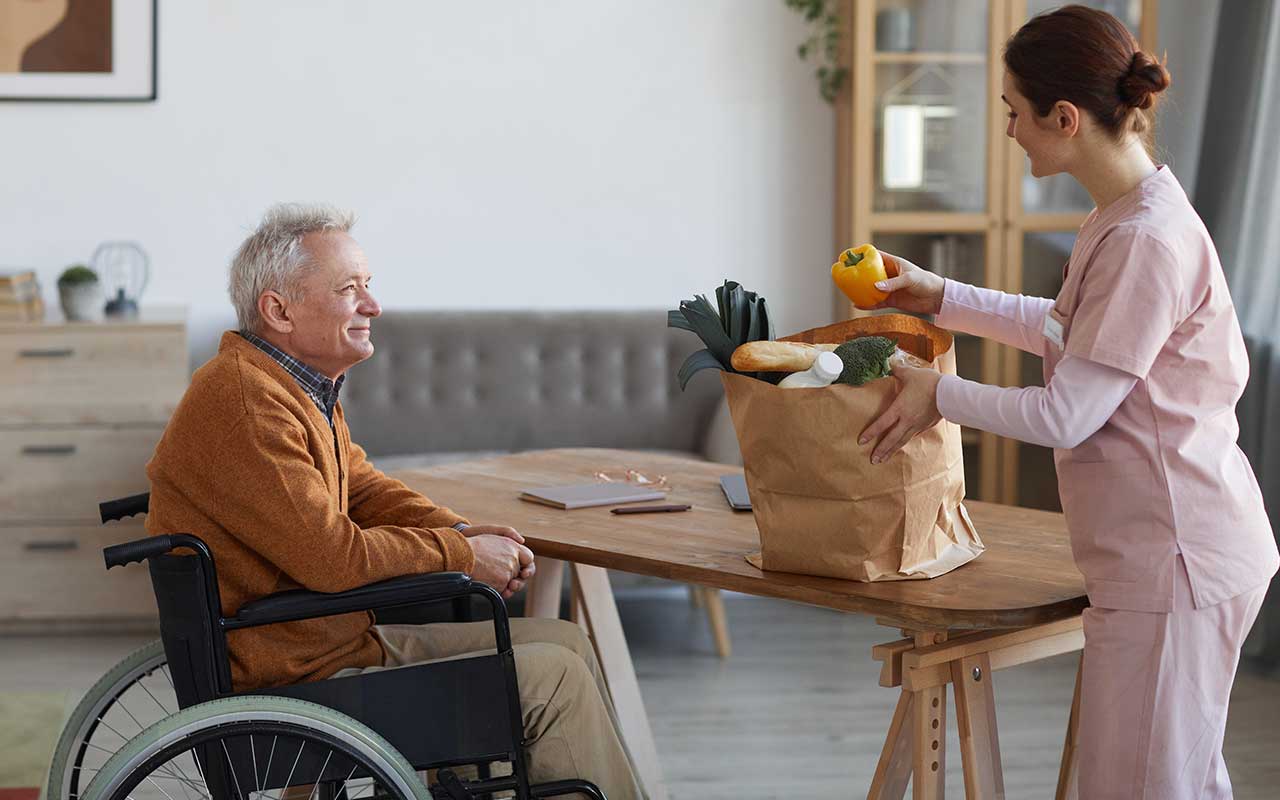 A man in a wheelchair sitting at a table while a caretaker holds a bag of groceries.