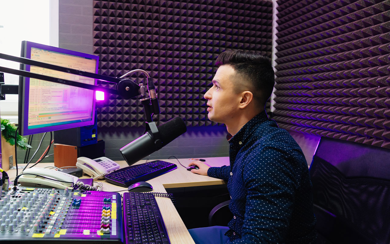 A man looking at a monitor, while talking into a microphone and using a sound board.