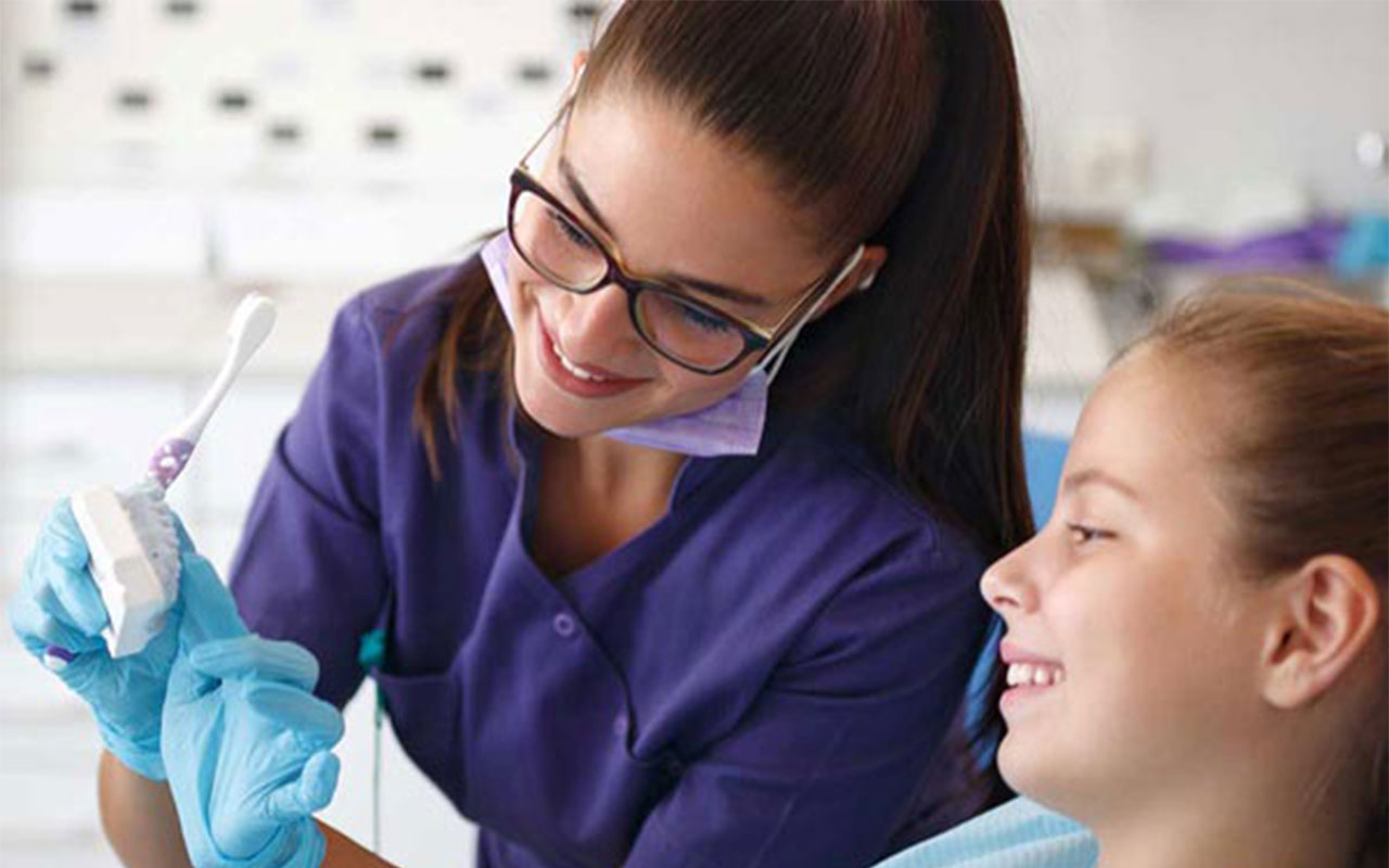 A woman holding a toothbrush and a mold of teeth, showing it to a little girl.