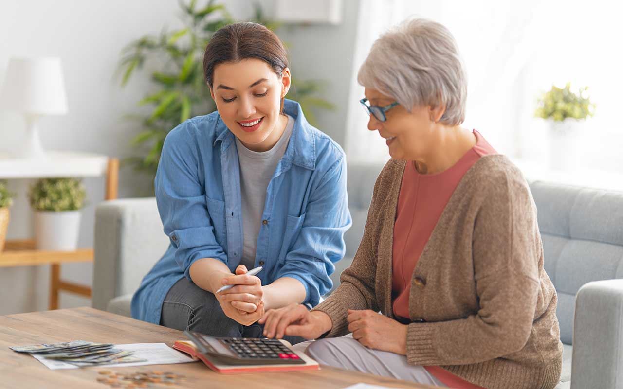 Two women using a calculator, sitting on a couch in a home.