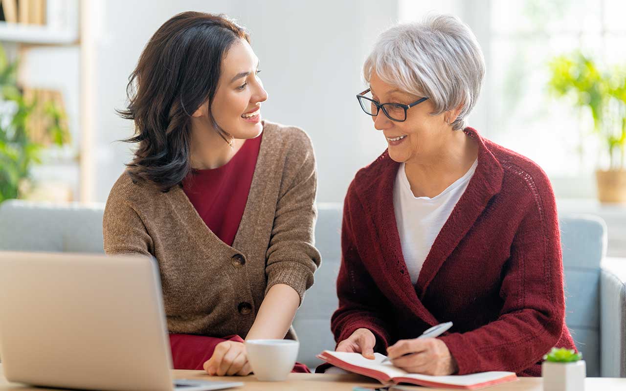 One woman referencing a laptop while the second lady writes in a notebook.