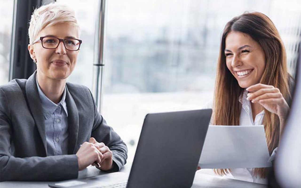 Two business women in an office with a laptop and papers, smiling