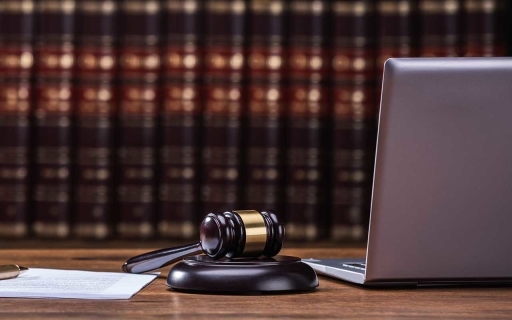 A gavel, pen, papers and laptop on a desk with legal books on a shelf behind.