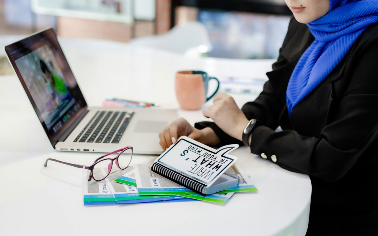 A business woman opening a notebook while sitting at a table with a laptop, glasses, coffee mug, pens and papers.