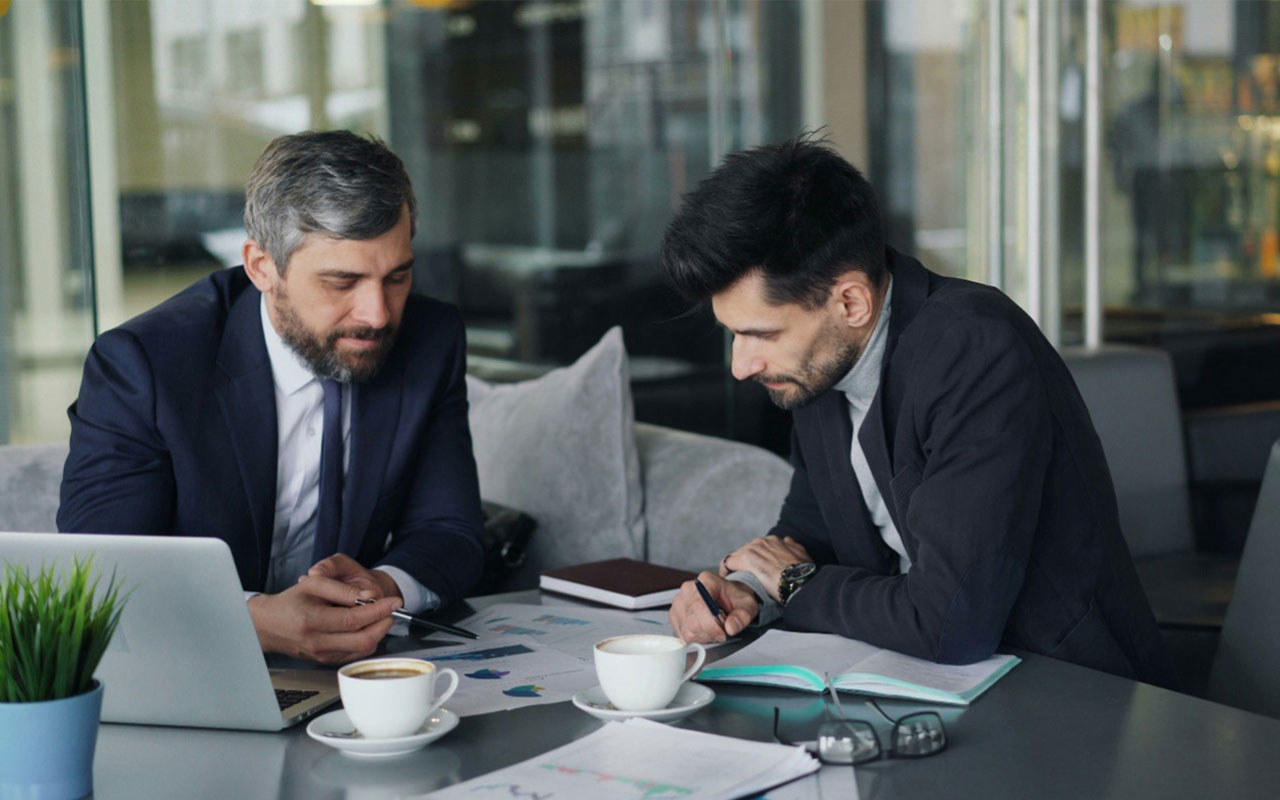 Two men in business suits holding pens, hunched over a table with open notebooks, coffee cups, papers, a pair of glasses and an open laptop.