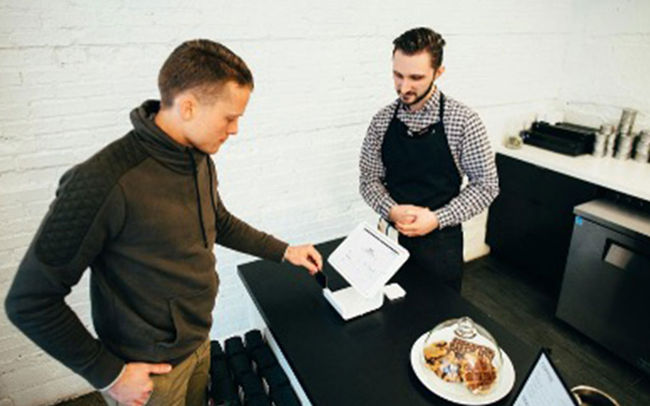 Two men in a cafe standing at a checkout counter. One man is in an apron and the other is swiping his credit card.