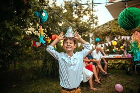 in glücklicher Junge feiert im Garten bei Tageslicht seinen Geburtstag, lacht und balanciert einen Partyhut auf seinem Kopf, während im Hintergrund Erwachsene an einem festlich dekorierten Tisch sitzen und den Moment genießen.