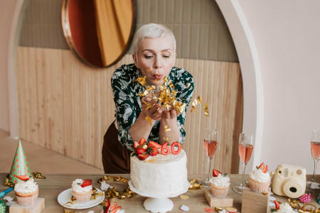 Frau pustet Konfetti von den Händen, feiert den 60. Geburtstag mit Kuchen und Sektgläsern im Hintergrund