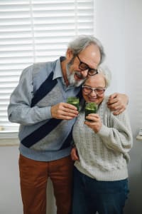 Ein lächelndes älteres Paar, das sich herzlich im Arm hält und gemeinsam grüne Smoothies in Gläsern hält