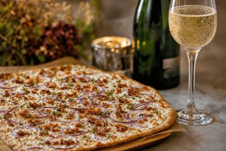 Knuspriger Flammkuchen mit Speck und roten Zwiebeln neben einem Glas Champagner auf einem Holzbrett.
