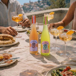 Zwei Flaschen Spritz-Getränk auf einem gedeckten Tisch mit Antipasti und Gläsern, während Menschen in sommerlicher Atmosphäre gemeinsam essen und trinken