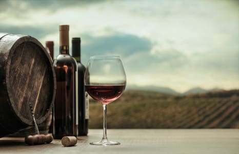 Glas Rotwein vor Weinflaschen und Holzfass mit Blick auf Weinberge in sanfter Abendstimmung.