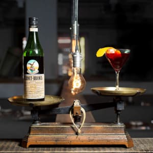 Altmodische Waage mit Fernet-Branca-Flasche in der linken und einem Martini-Cocktailglas in der rechten Schale.