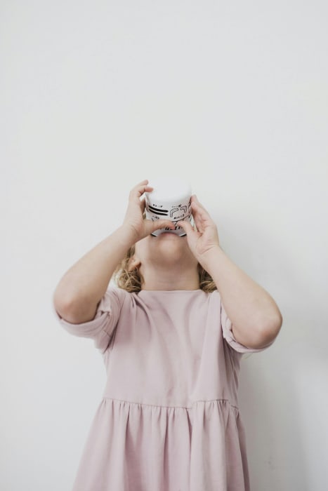 Ein kleines Mädchen in einem rosafarbenen Kleid hält eine große Tasse mit beiden Händen an ihren Mund und trinkt mit dem Kopf im Nacken
