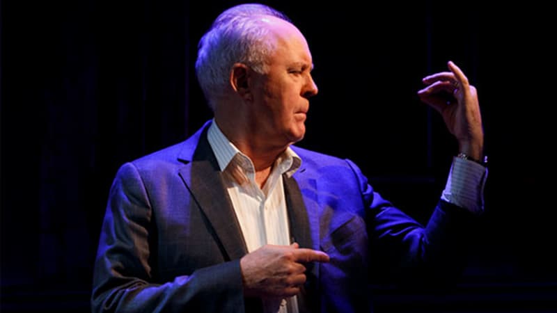 An older man in a white collared shirt and gray blazer holds up and examines his left hand while a wash of blue light showers him.