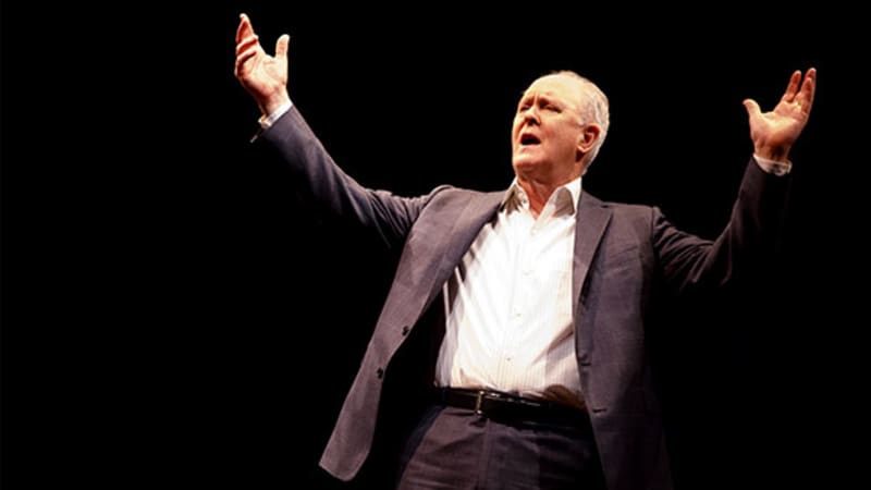 An older man in a white collared shirt and gray blazer stands with his arms up and out in a grand gesture.