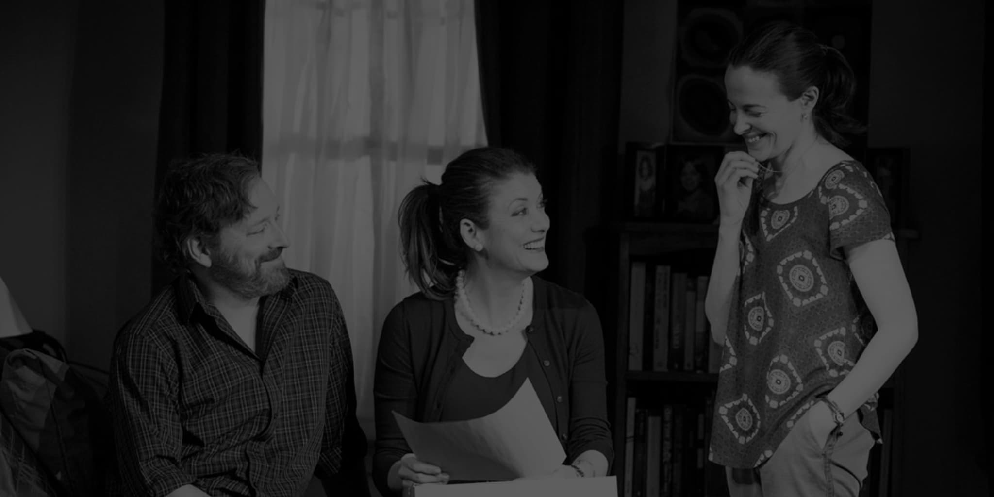 Production photo from IF I FORGET. A man and woman sit on the edge of a bed and a woman stands next to them. They all smile as the woman on the bed holds a box and a sheet of paper. 