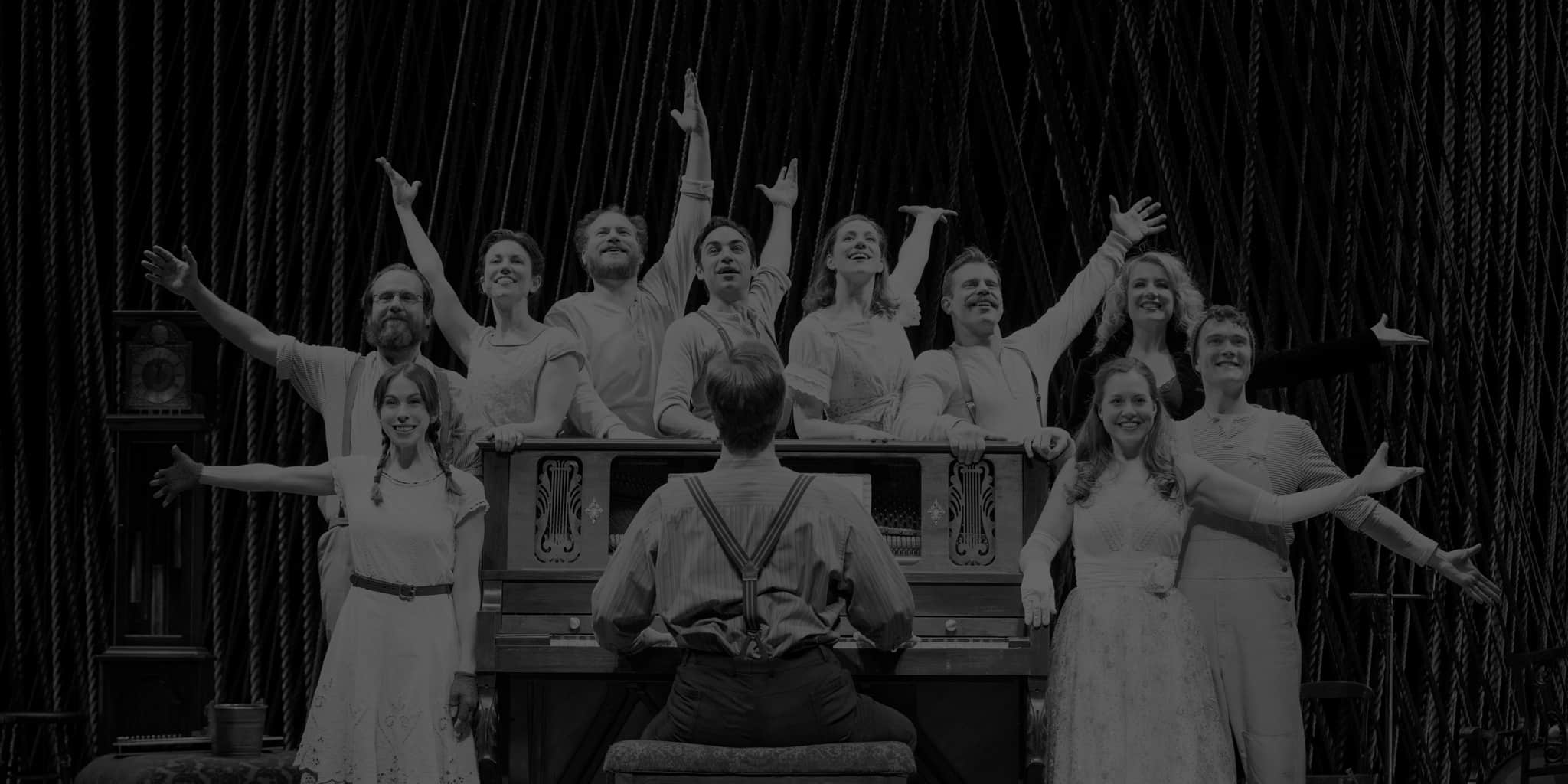 Production photo from INTO THE WOODS. The cast, all dressed in white, pose with their arms out and stand around a man playing the piano.