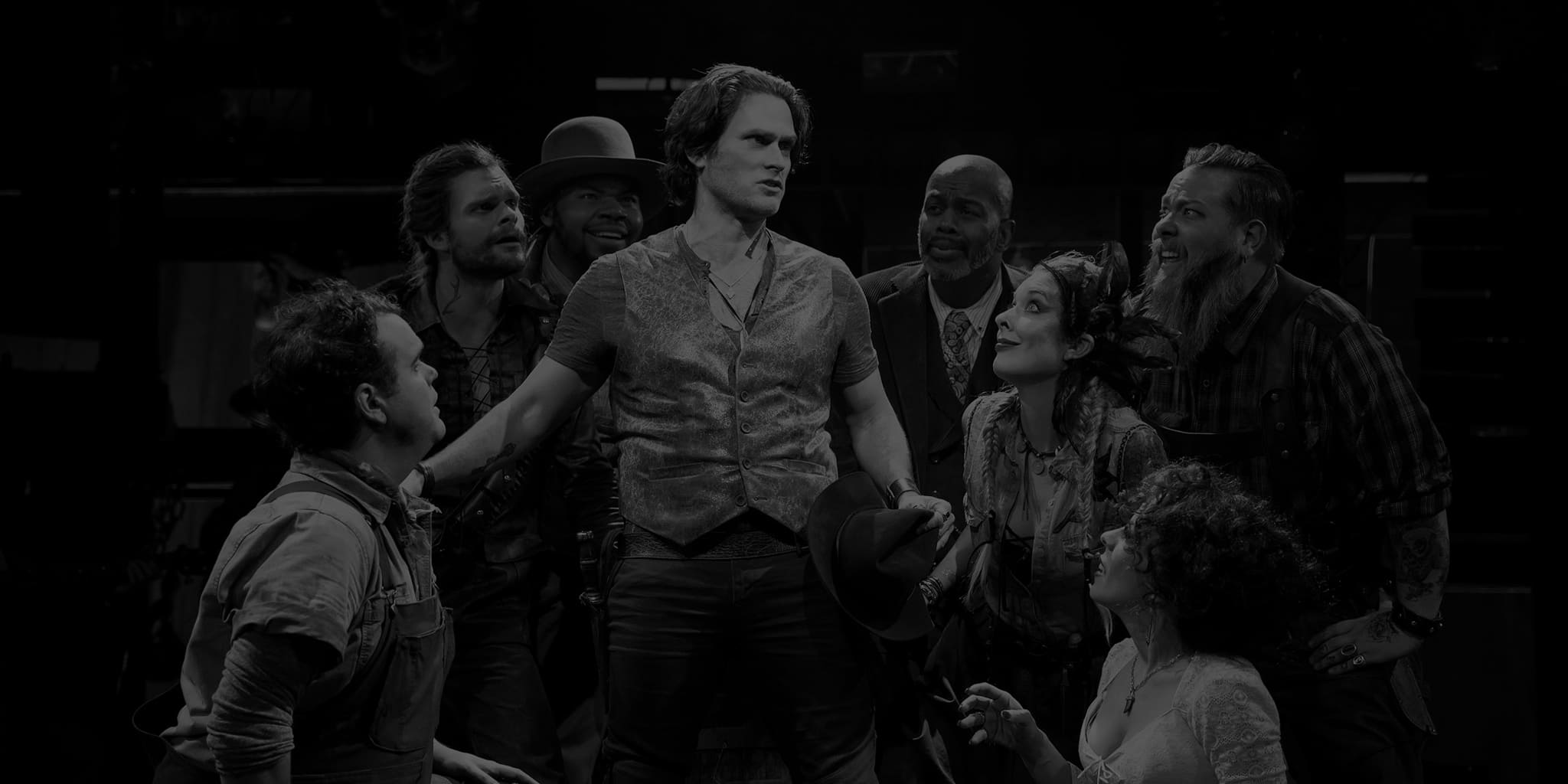 Production image from THE ROBBER BRIDEGROOM. A group of actors clump around a man wearing a vest over a t-shirt. He holds a hat in his hand and wears a pistol on his hip.