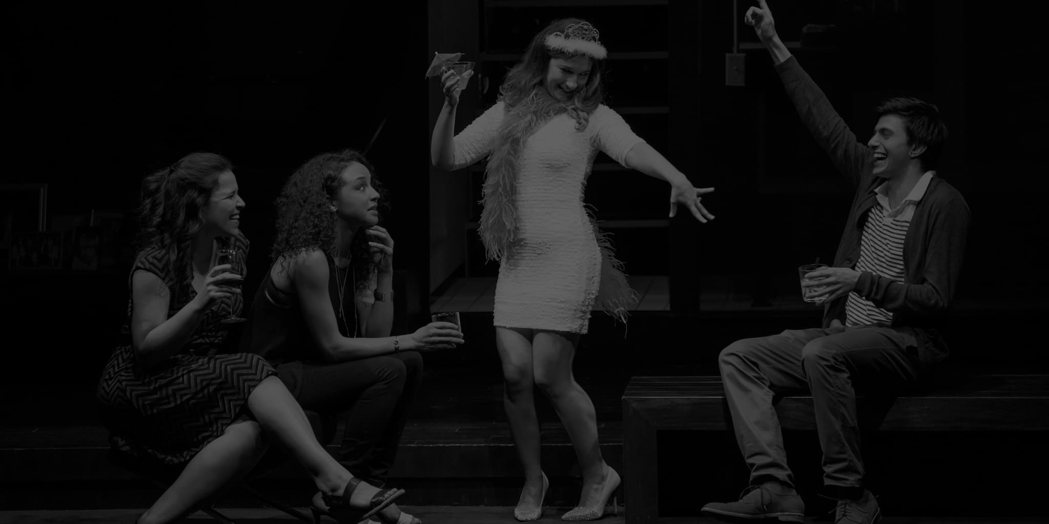 Production image from SIGNIFICANT OTHER. Three friends with drinks in hand sit around and hype up a dancing woman in a pink feather boa holding a tropical drink.