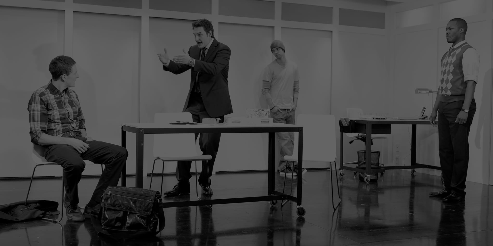 3 men in an office setting standing while one is yelling at a man sitting at a desk. 