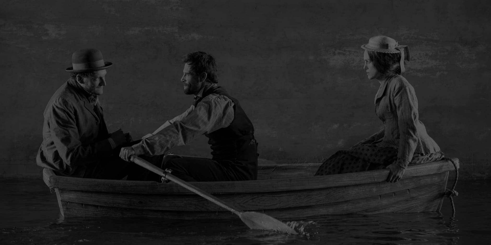 Production photo from THERESE RAQUIN. A man and woman in 1860s clothing sit on either ends of a wooden boat, while a man in the middle paddles them over a body of water.