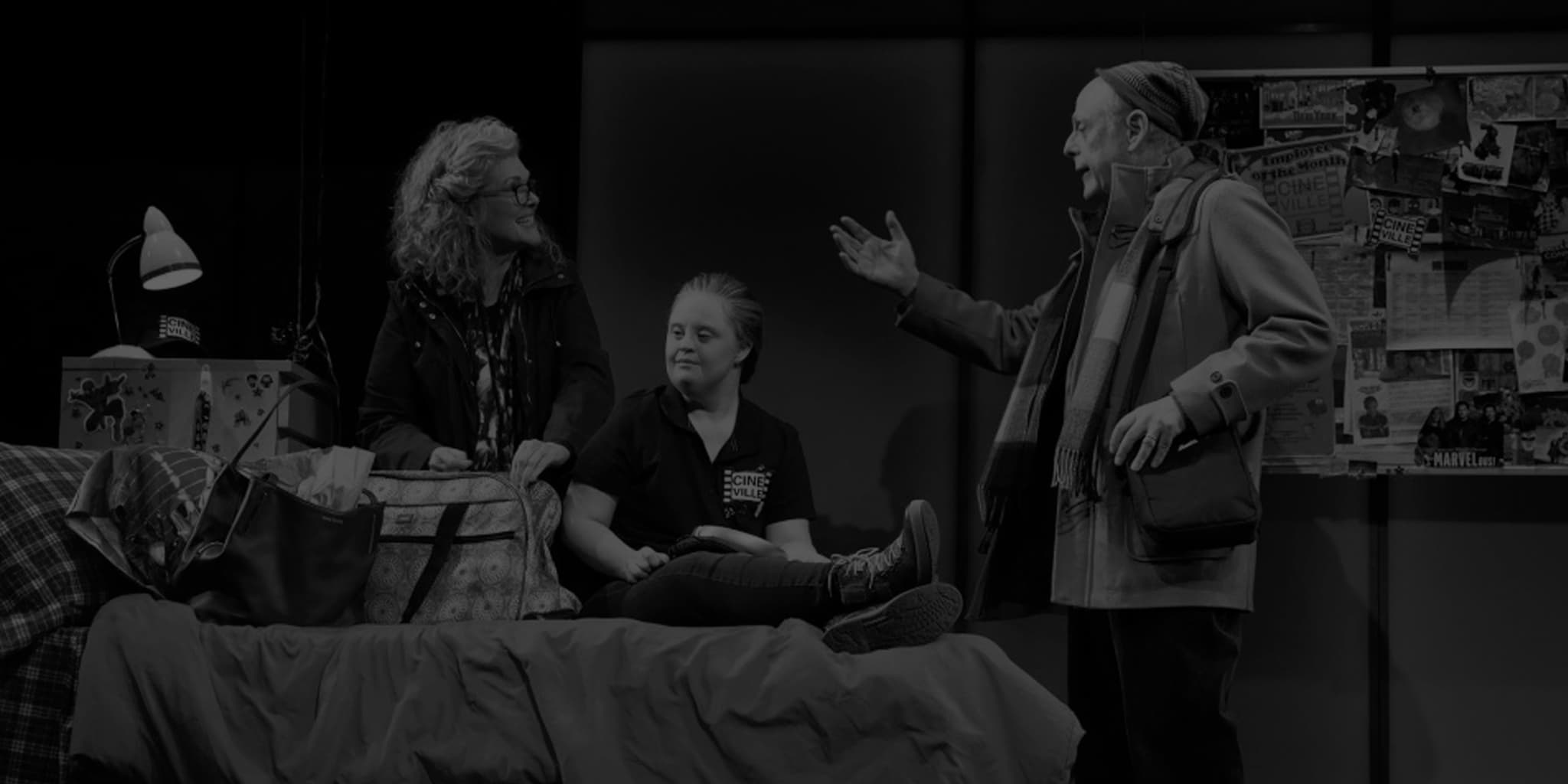 Production photo from Amy and the Orphans. A woman with Down syndrome sits on a bed full of bags while an older woman and man stand on either side of her in a heated conversation.