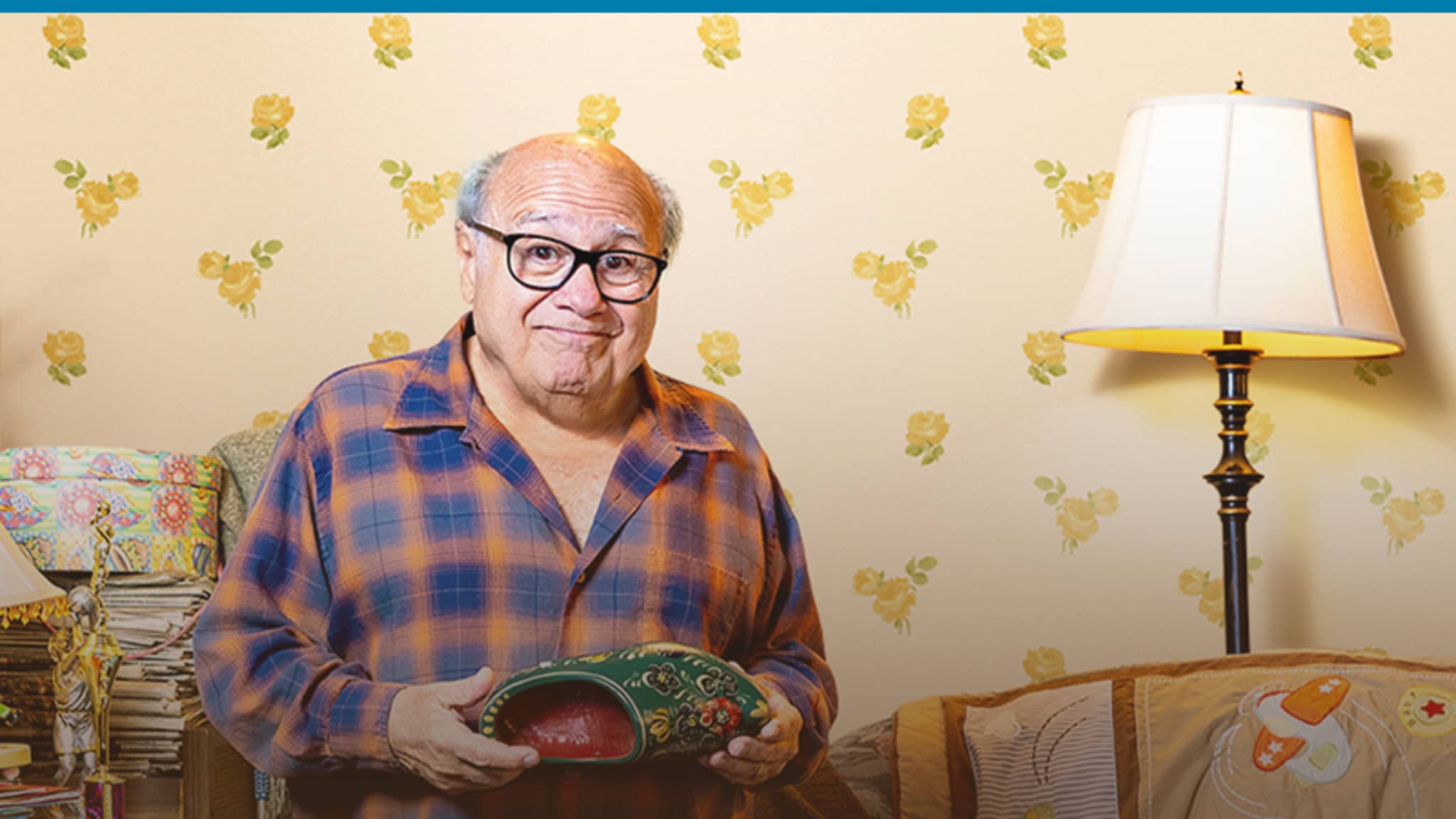 Danny DeVito wearing pajamas standing in a room filled with miscellaneous items all over the place.