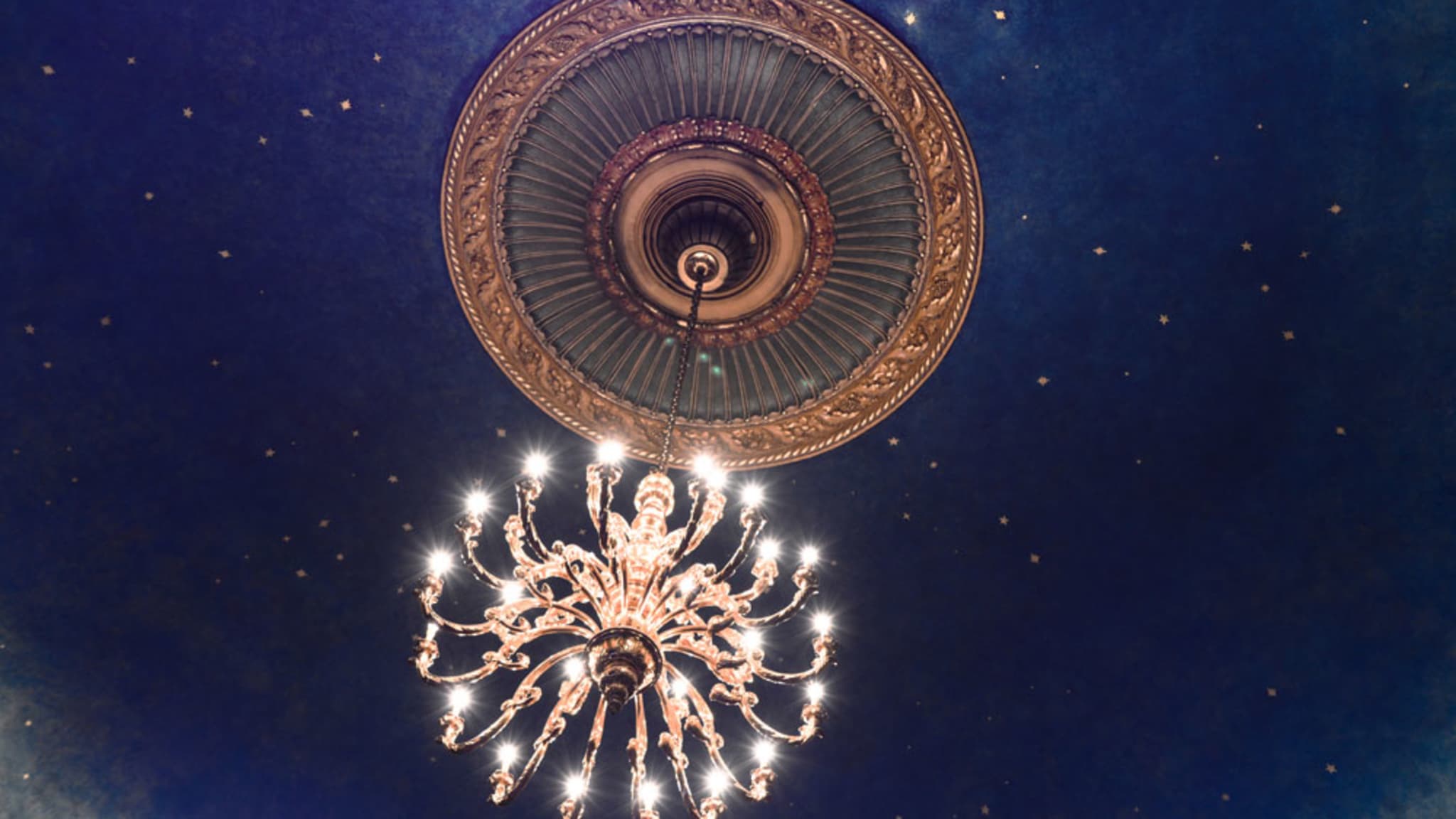 A large golden chandelier with bright white lights hangs from a decadent circle on a blue ceiling with sparkles and