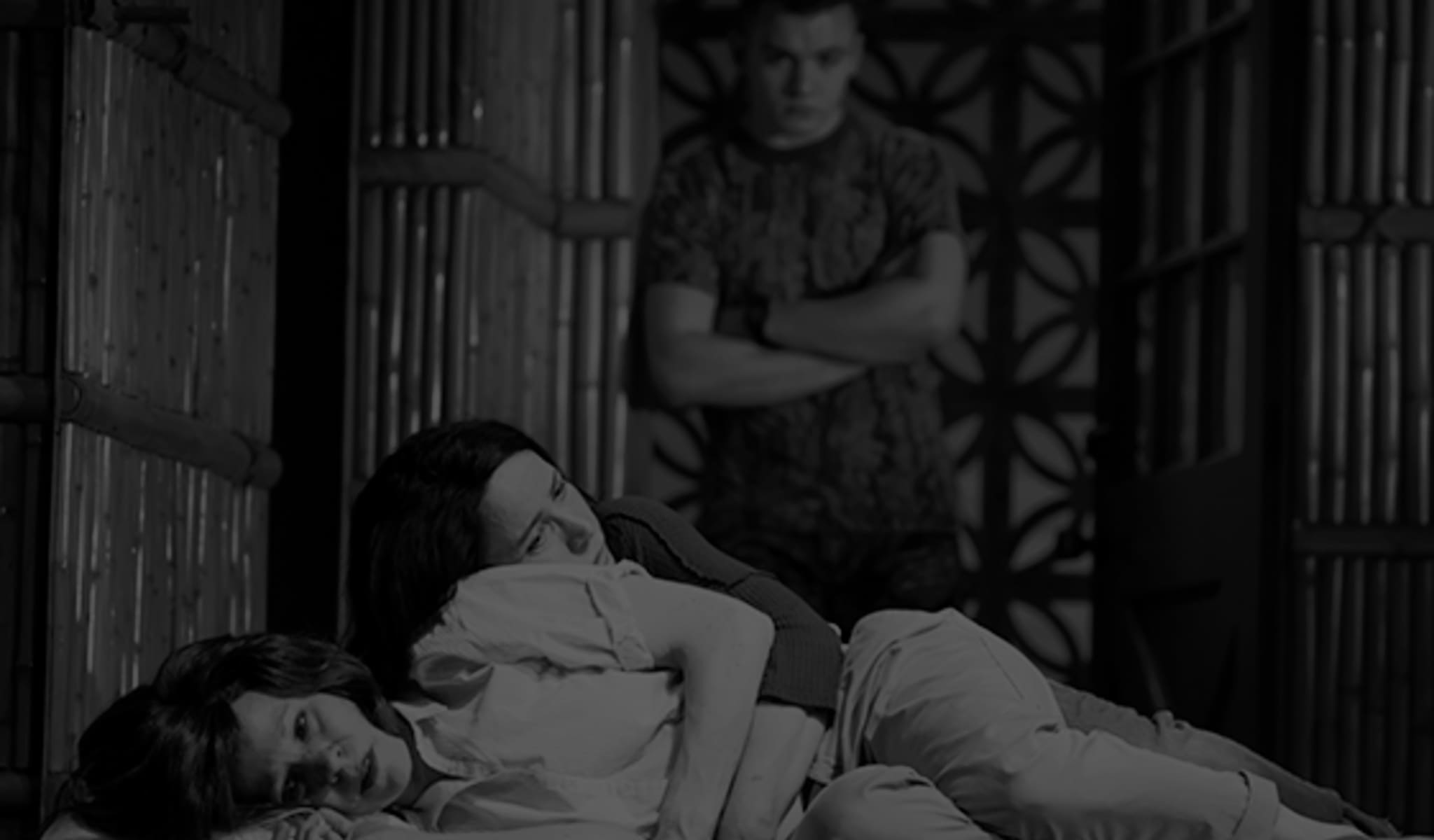 Production photo from MARVIN'S ROOM. Two upset women lay down and hold each other in a room with bamboo walls. A man crosses his arms and stands behind them.