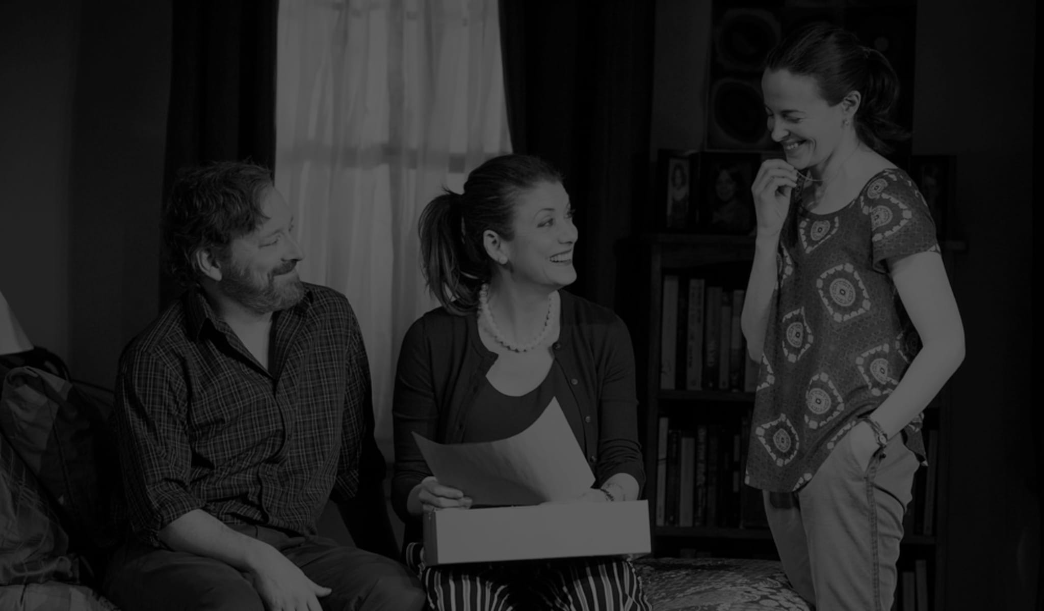 Production photo from IF I FORGET. A man and woman sit on the edge of a bed and a woman stands next to them. They all smile as the woman on the bed holds a box and a sheet of paper. 