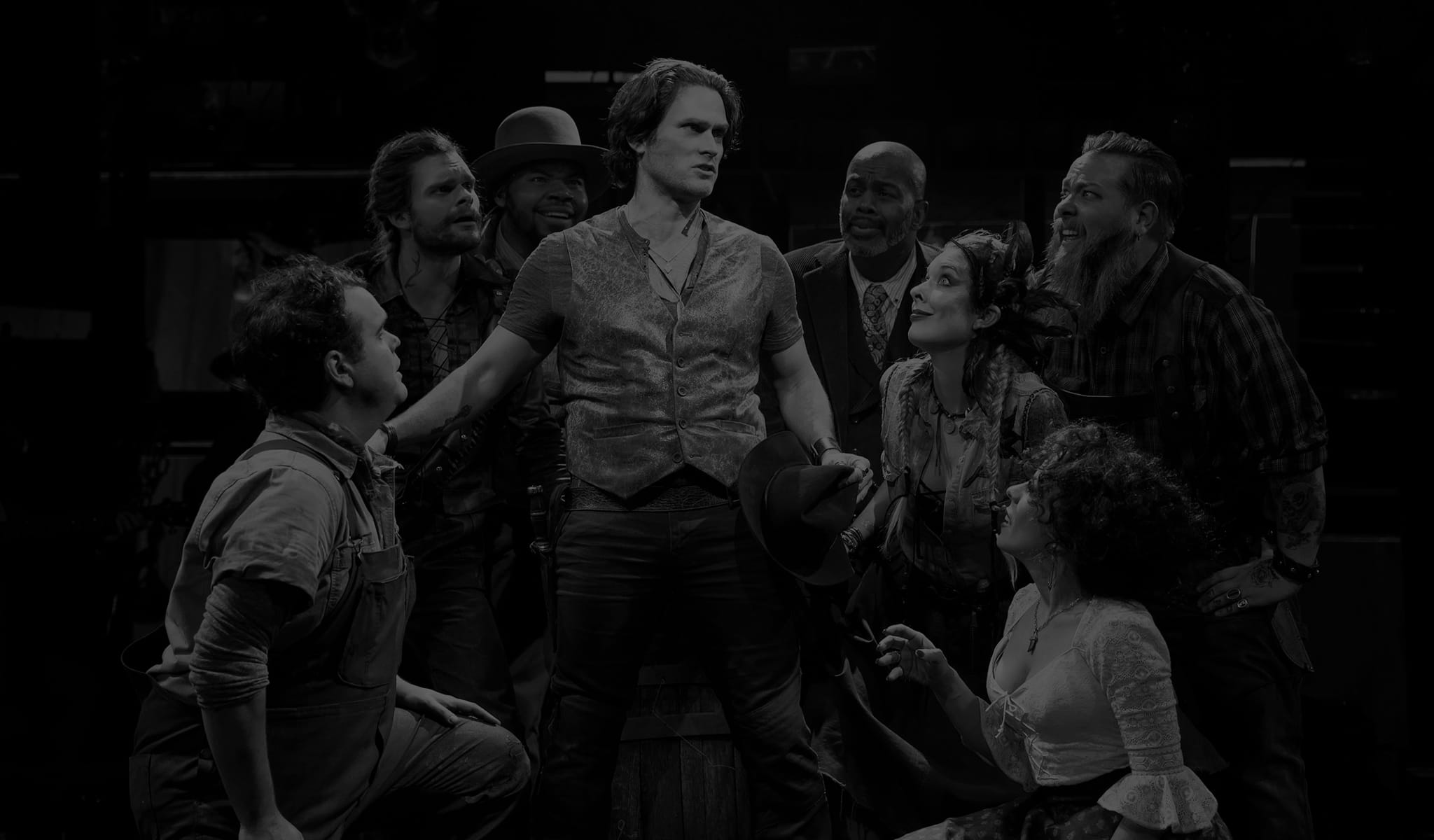 Production image from THE ROBBER BRIDEGROOM. A group of actors clump around a man wearing a vest over a t-shirt. He holds a hat in his hand and wears a pistol on his hip.