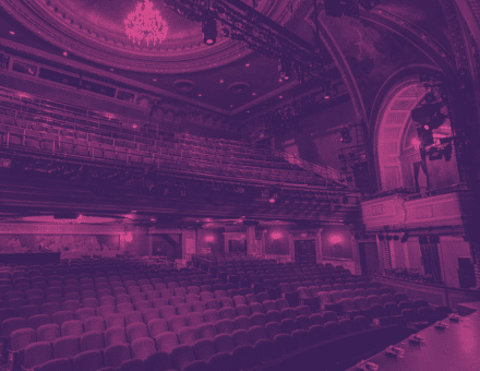 The empty house of the Todd Haimes Theatre with a wash of purple light over the seats, and a chandelier hangs at the top of the theatre.