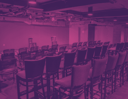 The empty house of Black Box Theatre. Two chairs are washed in red and yellow light at the front of rows and rows of chairs.