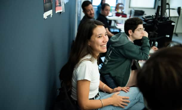 A row of people sit behind folding tables in the rehearsal studio, while one woman looks towards the camera and smiles.