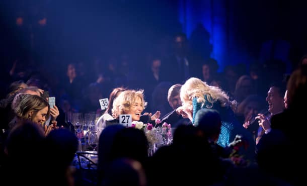 A spotlight on a table at a Roundabout gala. A woman with blonde hair smiles wide at another woman with a mic.