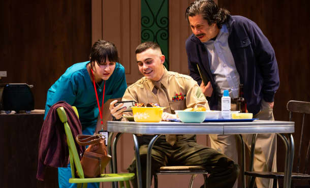 Two actors on stage look over the shoulder of an actor sitting at a breakfast table as he holds his iPhone horizontally. From 72 MILES TO GO.