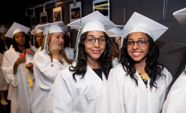 In a crowd of graduating students in white caps and gowns, two girls in their regalia smile at the camera.