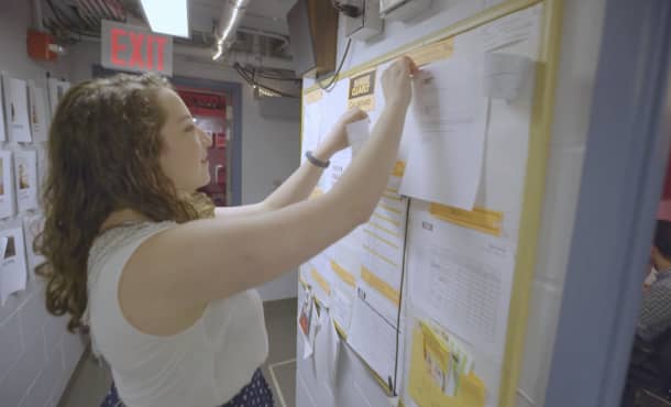 A young woman pins a piece of paper onto a cluttered bulletin board backstage.