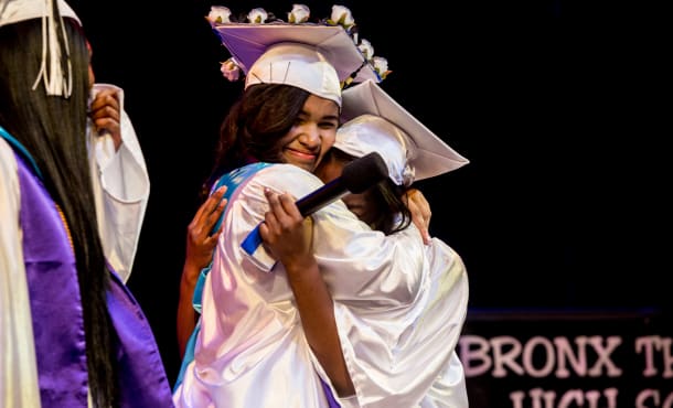 Two girls in white graduation regalia hug.