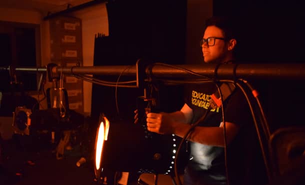 A student holds a spotlight hanging on a pole of other theatrical lamps and aims it away. An orange glow casts over their face.