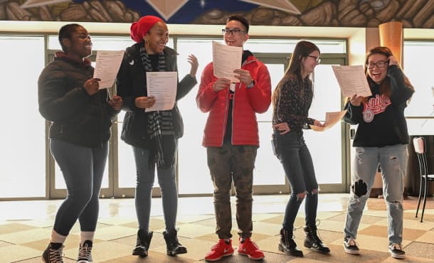 Five students stand in a line and hold lyric sheets as they laugh and sing in the American Airlines penthouse lobby.