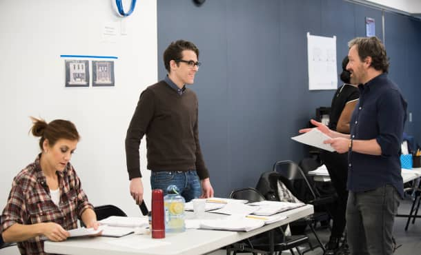 Two men stand across from each other in a rehearsal studio. Set sketches hang on the walls.